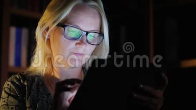 中年妇女工作到很晚，享受平板电脑。 平板电脑的屏幕反映在她的眼镜上